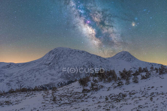 Breathtaking scenery of slope of hill covered with snow and trees against high rocky mountains under night starry sky with Milky Way — Stock Photo