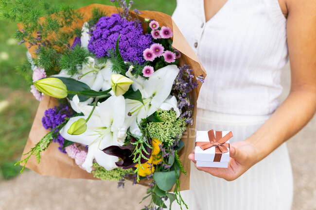 Crop anonymous female with small present box and blossoming flower bouquet in daytime on blurred background — Stock Photo