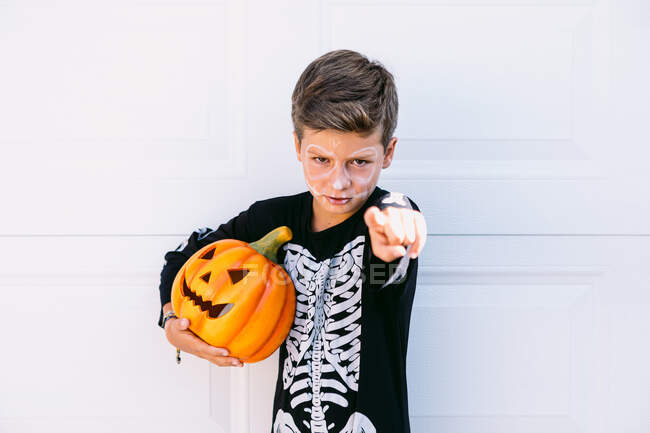 Серьёзный мальчик в костюме чёрного скелета держит тыкву Джека О Фонаря на Хэллоуин и указывает на камеру, стоя на белом фоне — стоковое фото