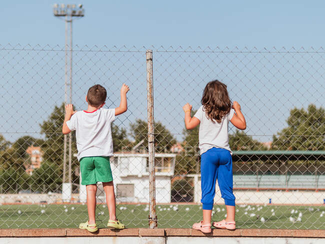 Back view full length of anonymous kids grabbing metal net fencing football field in stadium — Stock Photo