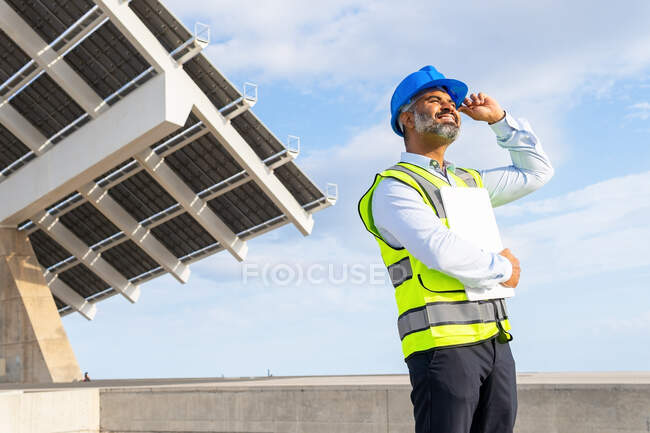 Low angle of mature ethnic male inspector in vest and hardhat with clipboard looking away while standing near solar power station — Stock Photo