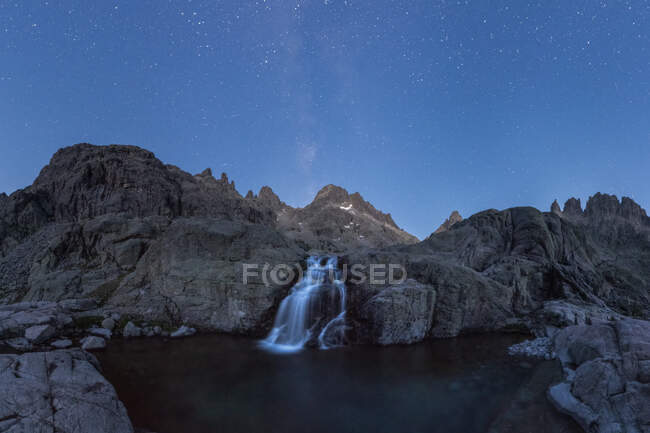 Espectacular paisaje de formaciones rocosas ásperas con cascada que fluye hacia el lago bajo cielo estrellado sin nubes por la noche - foto de stock