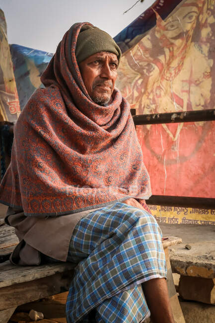 INDIA, VARANASI - NOVEMBER 27, 2015: Ethnic thoughtful male sittinglooking away on wooden bench on shabby village in India — Stock Photo