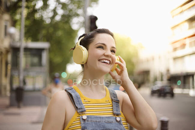 Mujer joven y positiva escuchando música en auriculares inalámbricos mirando a la cámara mientras camina por la calle - foto de stock