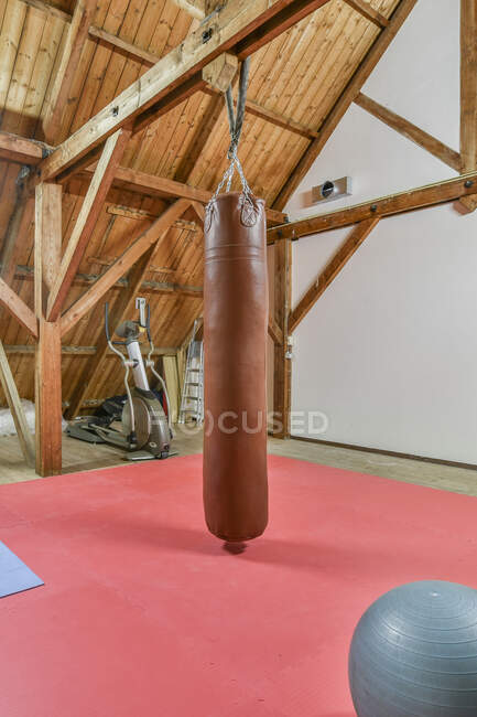 Punching bag hanging between fitness ball and elliptical trainer in attic above mat at home — Stock Photo