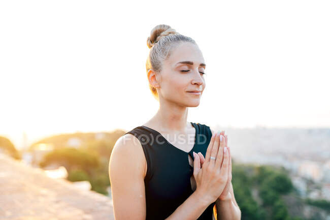 Спокойная женщина с руками на груди и закрытыми глазами медитирует на крыше во время занятий йогой вечером — стоковое фото