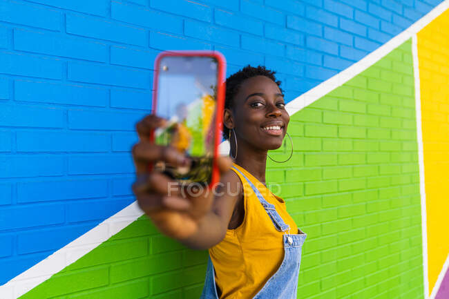 Selective focus of cellphone in hands of cheerful African American female taking self portrait against bright wall — Stock Photo