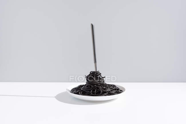 Stainless fork in full bowl of black spaghetti with cuttlefish ink on white table in studio on gray background — Stock Photo