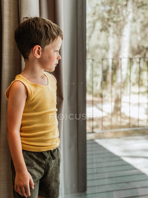 Petit enfant serein debout près de la fenêtre dans le chalet et regardant loin dans les pensées — Photo de stock