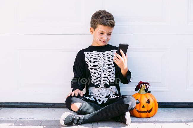 Ganzkörper fröhlicher Junge in schwarzem Skelettkostüm mit bemaltem Gesicht und geschnitztem Halloween-Kürbis, der auf dem Handy sitzt, während er in der Nähe der weißen Mauer auf der Straße sitzt — Stockfoto