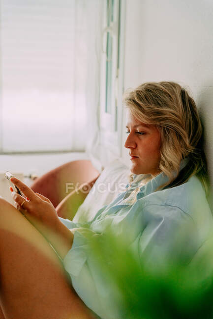 Vista lateral de la hembra adulta en camisa navegando por Internet en el teléfono móvil mientras está sentado en la habitación de luz - foto de stock