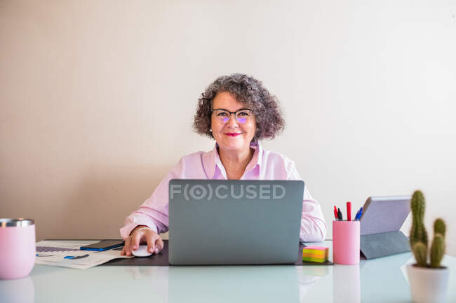 Весела старша бізнес-леді в окулярах сидить за столом з нетбуком і мишкою, дивлячись на камеру на світлому фоні — стокове фото