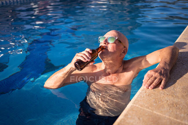 Angolo alto di rilassato maschio anziano in occhiali da sole bere birra dalla bottiglia di vetro mentre nuota in piscina — Foto stock