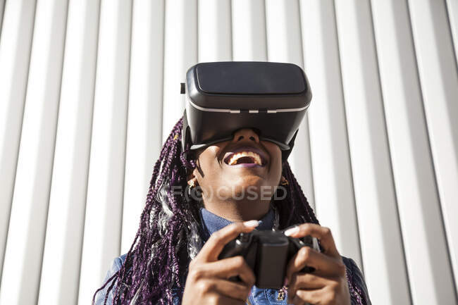 Задоволена молода афроамериканська жінка в VR-хедсеті з використанням контролера під час розваги і гри в віртуальну гру проти сірої смугастої стіни — стокове фото