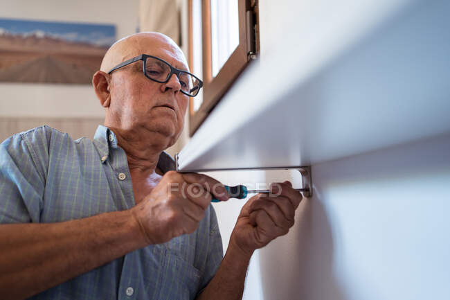 Hombre de edad avanzada atento en gafas con destornillador manual atornillar estante a la pared en la habitación de la casa - foto de stock