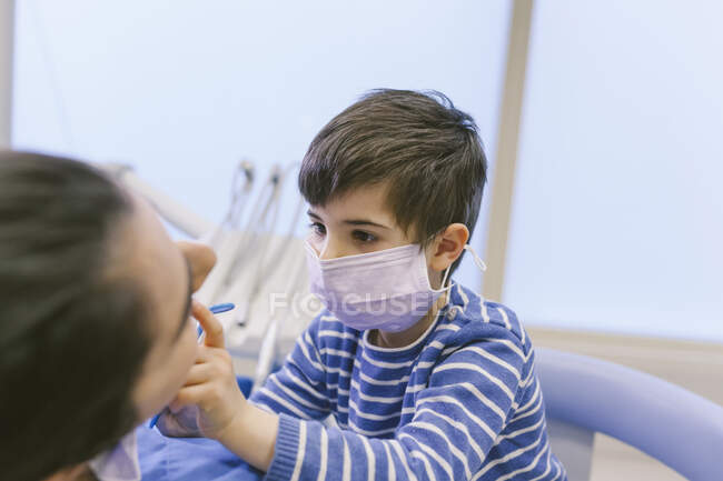 Цікавий хлопчик в медичній масці грає роль стоматолога і перевіряє зуби з дзеркалом в лікарні — стокове фото