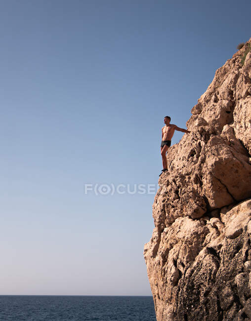 Turista masculino sin miedo en bañador contemplando mar con horizonte desde montaña agitada en Ibiza España - foto de stock