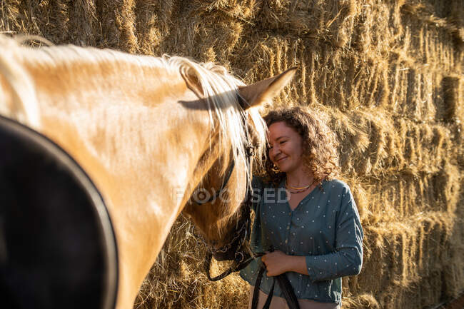 Content mature female stroking stallion muzzle with smooth brown coat against hay in riding school on sunny day — Stock Photo