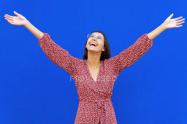 Positive young female with brown hair in red dress standing with raised arms and looking up against blue background in daytime — Stock Photo