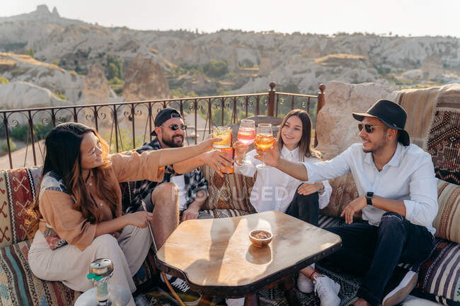 Высокий угол улыбающихся друзей, сидящих за столом и поднимающих бокалы с коктейлями, пока они тусуются в баре на террасе в Каппадокии, Турция — стоковое фото