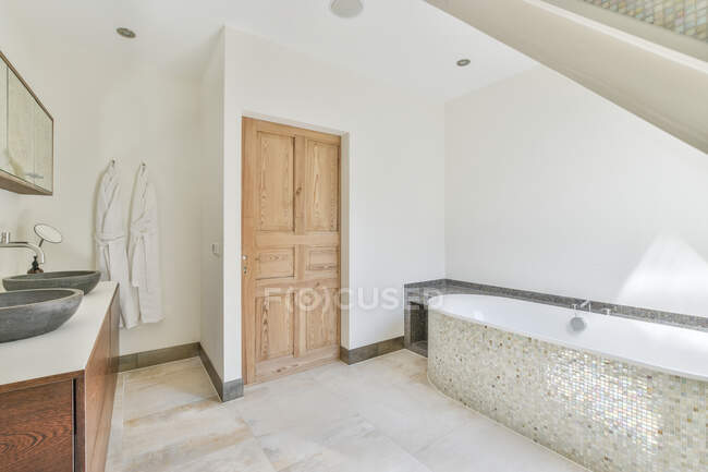 Bathtub with mosaic tiles and washbasins between cabinets and mirrors in contemporary bathroom on sunny day — Stock Photo