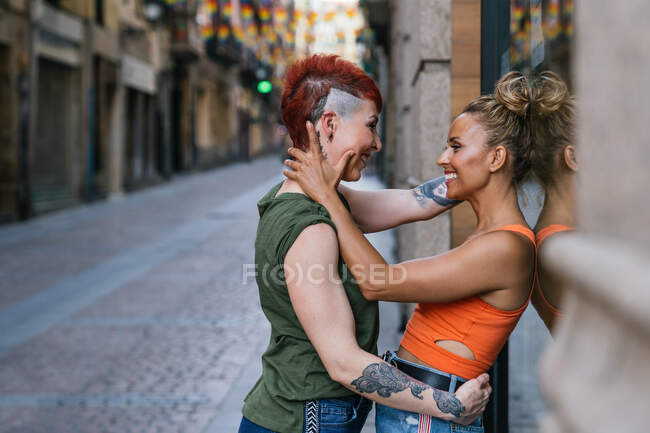 Vista laterale della giovane coppia lesbica allegra alla moda con tatuaggio che si abbraccia guardandosi in un momento di bacio appoggiato a un muro in città — Foto stock