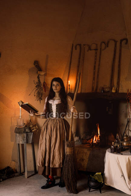 Hexe im langen Kleid, die mit einem Zauberbuch in die Kamera blickt, während sie in einem gemütlichen Raum mit Besen und Hexenkessel steht — Stockfoto