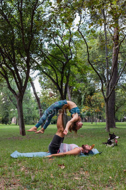 Vista laterale di coppia flessibile in abbigliamento sportivo praticare acroyoga insieme su tappetino yoga sull'erba contro gli alberi nel parco durante il giorno — Foto stock
