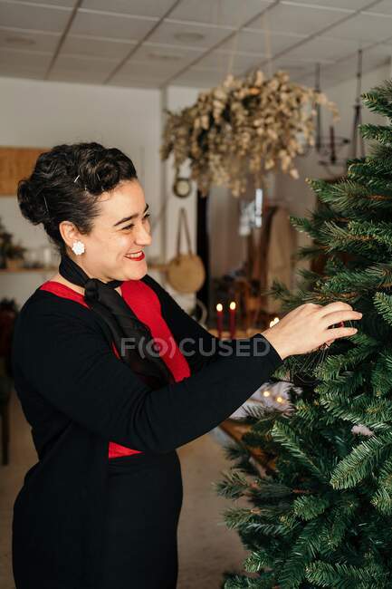 Вид збоку весела жінка з зачіскою в стильному одязі, що стоїть і прикрашає різдвяну ялинку в квартирі вдень — стокове фото
