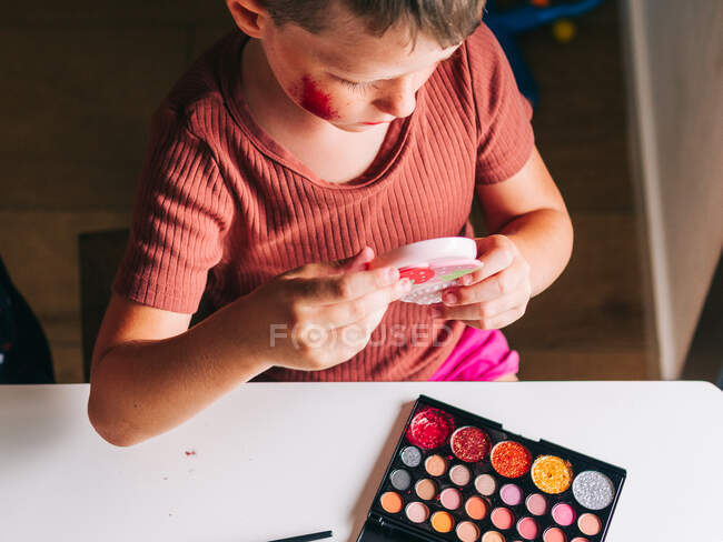 De cima da criança de colheita com maquiagem no rosto olhando no espelho à mesa com paleta de sombra em casa — Fotografia de Stock