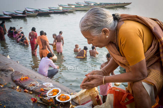 INDIA, VARANASI - NOVEMBER 27, 2015: Elderly ethnic woman praying and doing offers with candles and flowers near river in India — Stock Photo