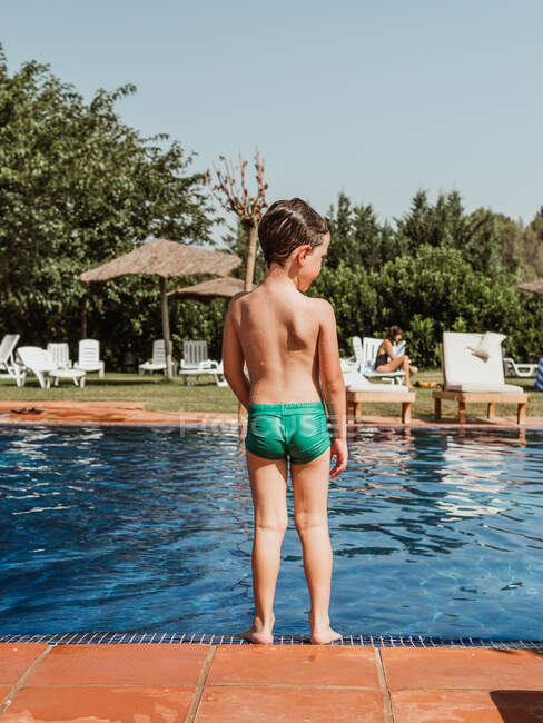 Back view of unrecognizable wet little boy in shorts standing near swimming pool on sunny day in summer — Stock Photo