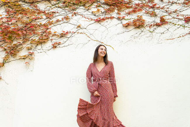 Mulher positiva em vestido elegante e óculos de pé na parede branca com ramos de planta olhando para longe durante o dia — Fotografia de Stock