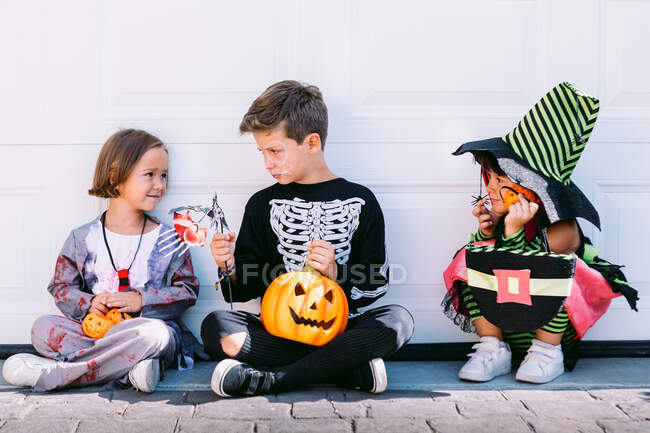 Cuerpo completo de grupo de niños pequeños vestidos con varios disfraces de Halloween con linterna Jack O tallada sentada cerca de la pared blanca en la calle - foto de stock