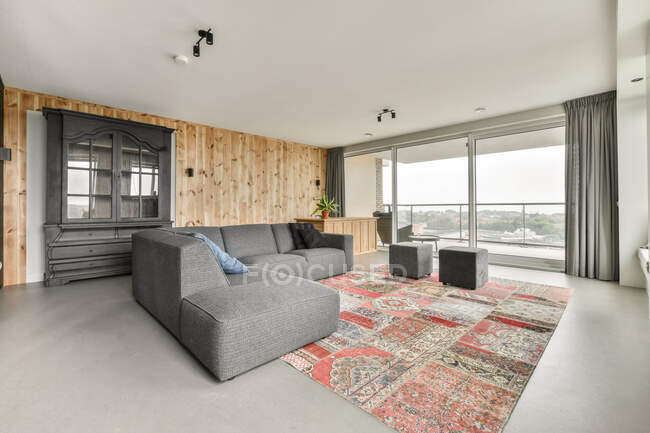 Divano grigio posizionato su tappeto colorato in soggiorno con pareti in legno e finestre panoramiche in appartamento moderno — Foto stock