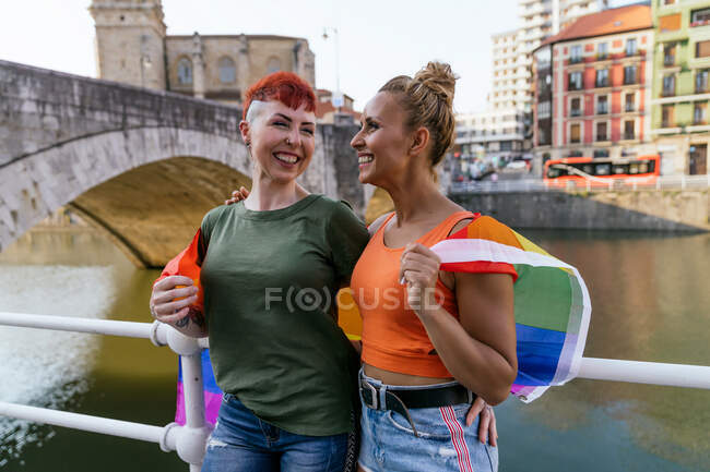 Cool mujer tatuada con mohawk y bandera LGBTQ abrazando novia con los ojos cerrados contra el canal en la ciudad - foto de stock