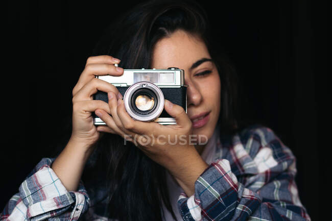 Concentré jeune femme avec les yeux fermés en appuyant sur le bouton tout en prenant des photos sur caméra vintage sur fond noir — Photo de stock