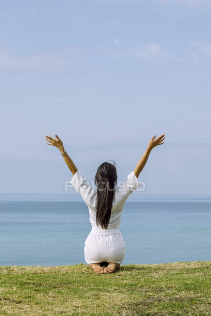 Back view of anonymous female with raised arms sitting on knees during yoga practice on green coast against ocean — Stock Photo