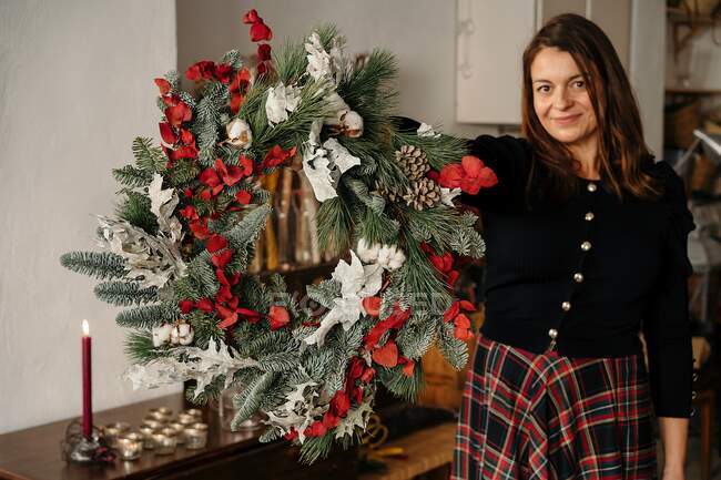 Весела жінка в повсякденному одязі стоїть з декоративним різдвяним вінком в руках дивлячись на камеру в кімнаті — стокове фото