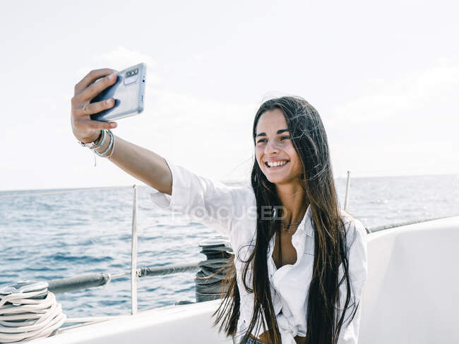 Adolescente sonriente de pelo largo tomando autorretrato en celular en lancha amarrada en el mar en Tenerife España - foto de stock