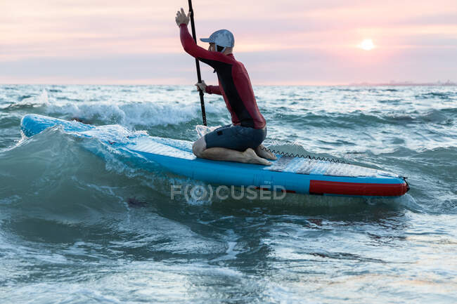 Вид збоку чоловічий серфер у гідрокостюмі та капелюсі на дошці для весла, що серфінгу на морському узбережжі — стокове фото