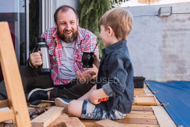 Niño sincero con calabaza calabaza de bebida infundida contra papá barbudo alegre con termos sobre fondo borroso - foto de stock