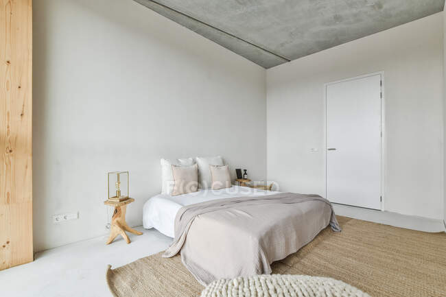 Креативный дизайн спальни с подушками и чехлом на кровати между дверью и столами на полу дома — стоковое фото