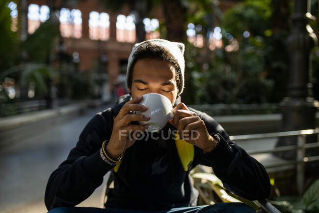 Homme afro-américain paisible hipster avec sac à dos froid assis avec les yeux fermés dans le parc et boire une boisson rafraîchissante à emporter — Photo de stock