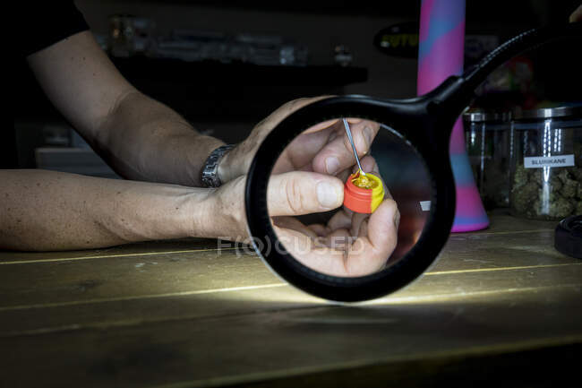 Ritaglia maschio irriconoscibile mostrando vaso con colla di marijuana e bastone attraverso lampada di ingrandimento nello spazio di lavoro — Foto stock