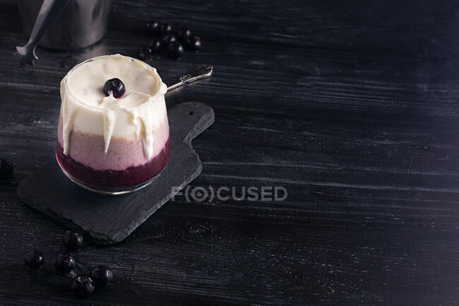 Verre de délicieux smoothie banane et myrtille avec crème fouettée sur planche à découper contre pot de yaourt et seau en métal — Photo de stock
