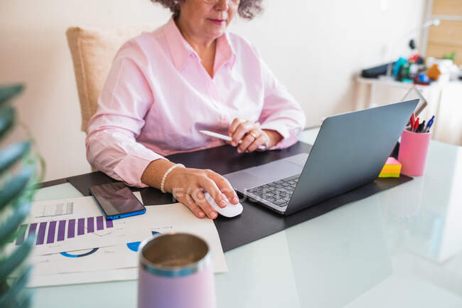 Crop senior female entrepreneur with tablet and netbook working at desk with graphics on paper sheets — Stock Photo