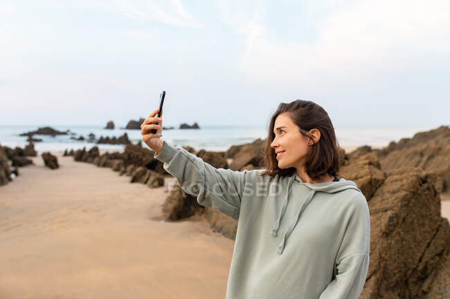 Friendly female taking self portrait on cellphone on sandy shore against rocks and ocean under cloudy sky in Asturias Spain — Stock Photo