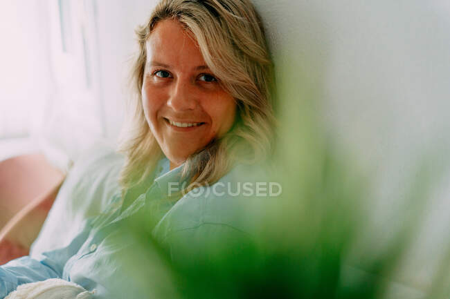 Friendly adult female with wavy blond hair looking at camera while sitting on bed in house — Stock Photo