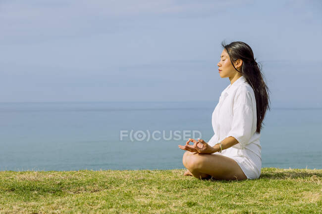 Vista lateral de la soñadora joven étnica femenina con los ojos cerrados meditando en Padmasana posando mientras practica yoga en la costa del mar - foto de stock